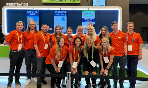 The Perenso team wearing orange polos and posing in front of a registration desk for a photo at a UNFI food show.